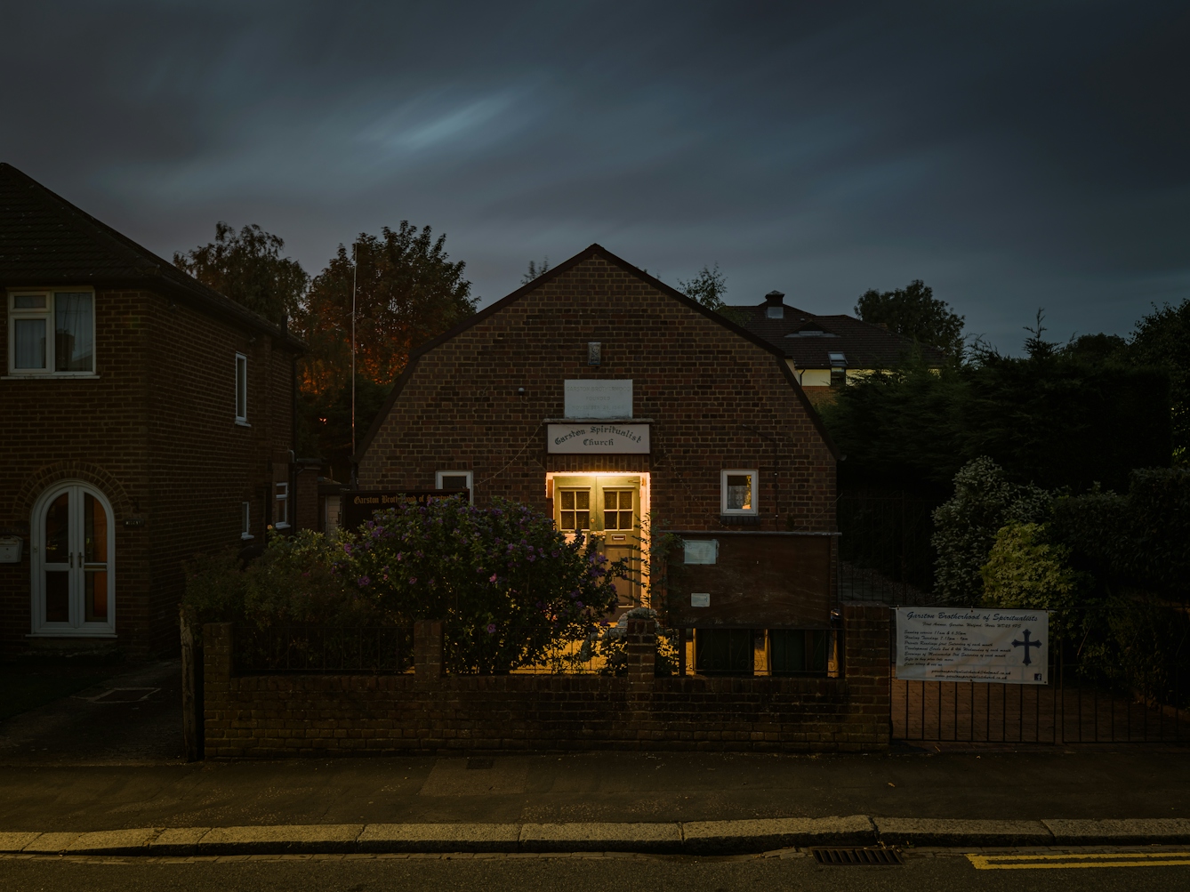 Photograph of Garston Spiritualist Church, Watford, at night.  Set in a residential setting the double doors of the church are lit by a porch light, which is also spilling into the small yard.  The church is partially hidden by a brick wall come gate, and two bushes. 