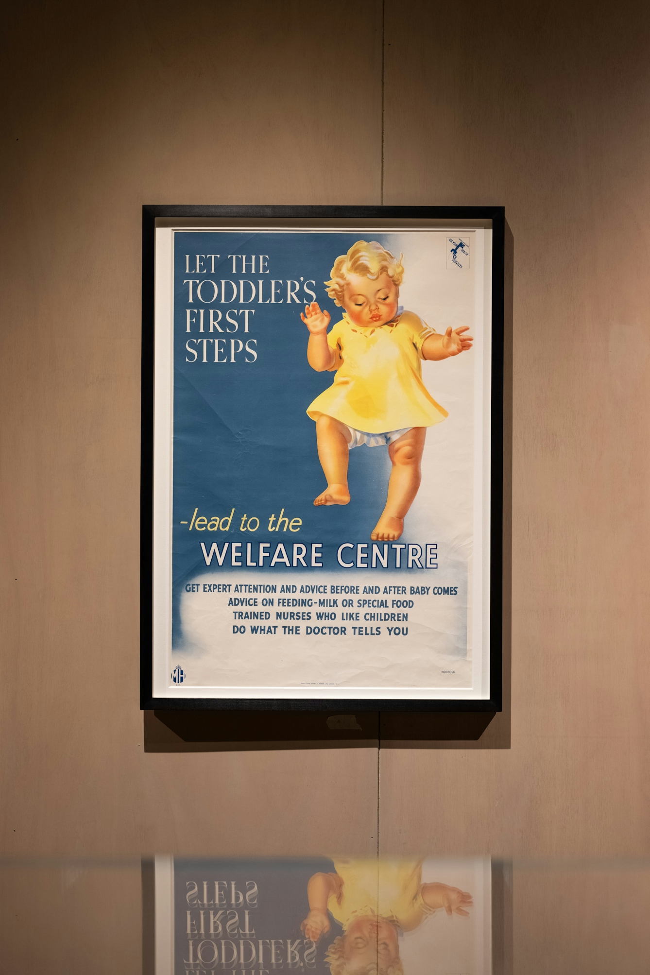 Photograph of a framed poster hanging in a frame on the wall of an exhibition gallery. The poster shows a blond haired toddler in a yellow dress. Beside them are the words, 'let the toddler's first steps - lead to the welfare centre'. 