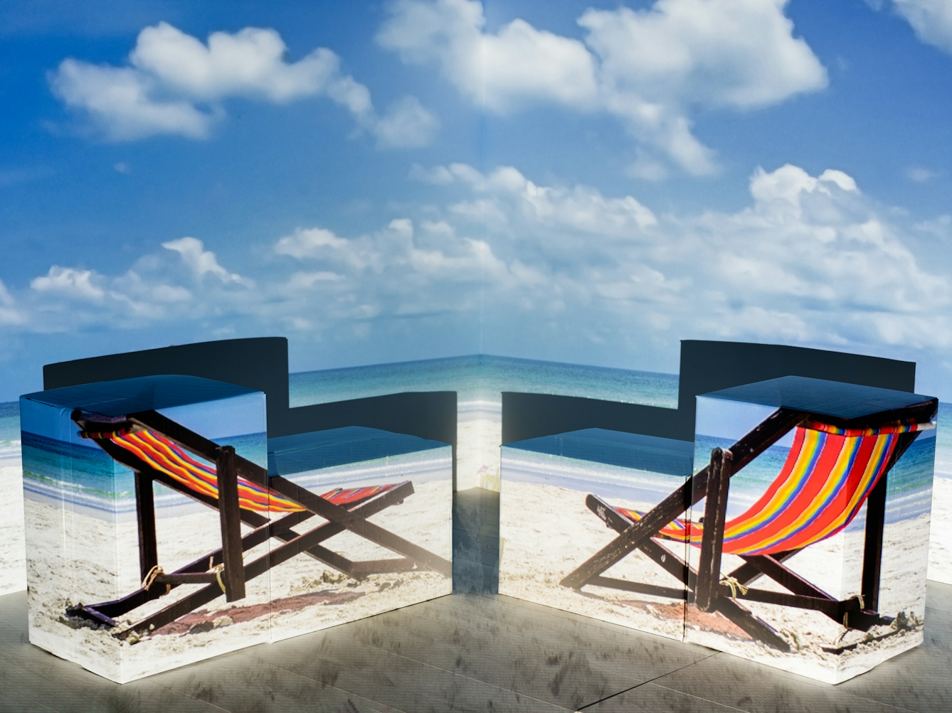 Photograph of the corner of a room showing the walls and a section of the wooden floor. On the floor is an arrangement of cardboard boxes. Onto the whole scene is projected an image of a beach with two stripy deckchairs. The boxes have assumed the shape of the deckchairs and have the images of them projected onto their sides.