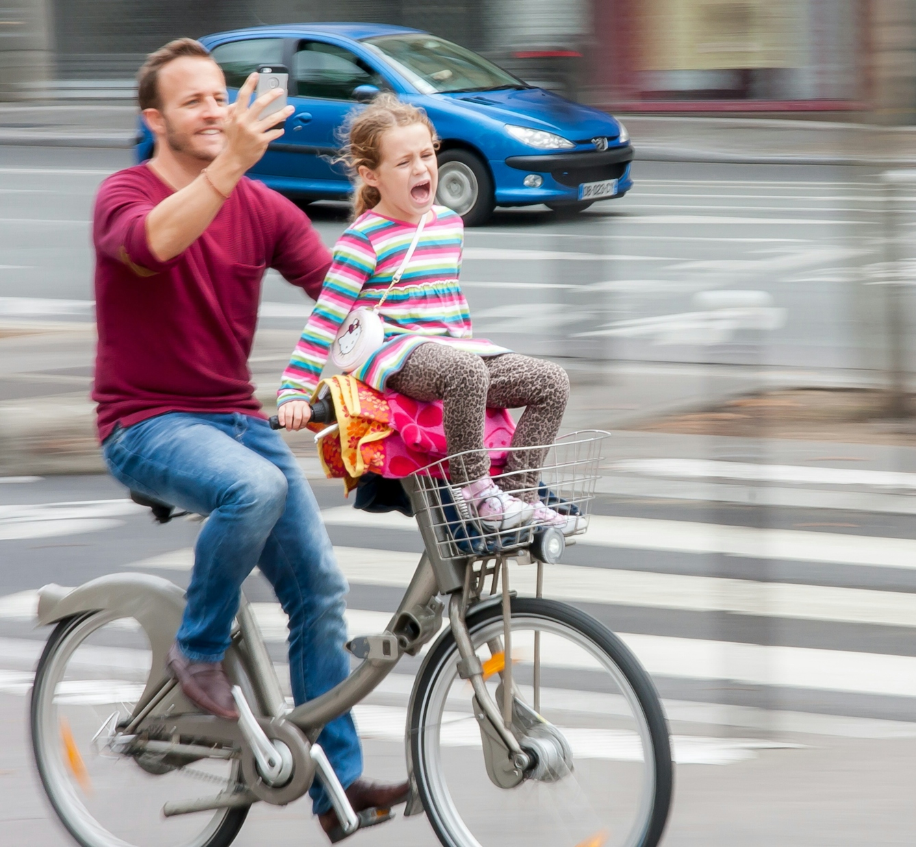 Photograph of a man riding a bicycle taking a selfie whilst the girl sitting on his handlebars appears to be crying or screaming. Blurred background and car behind indicate movement.
