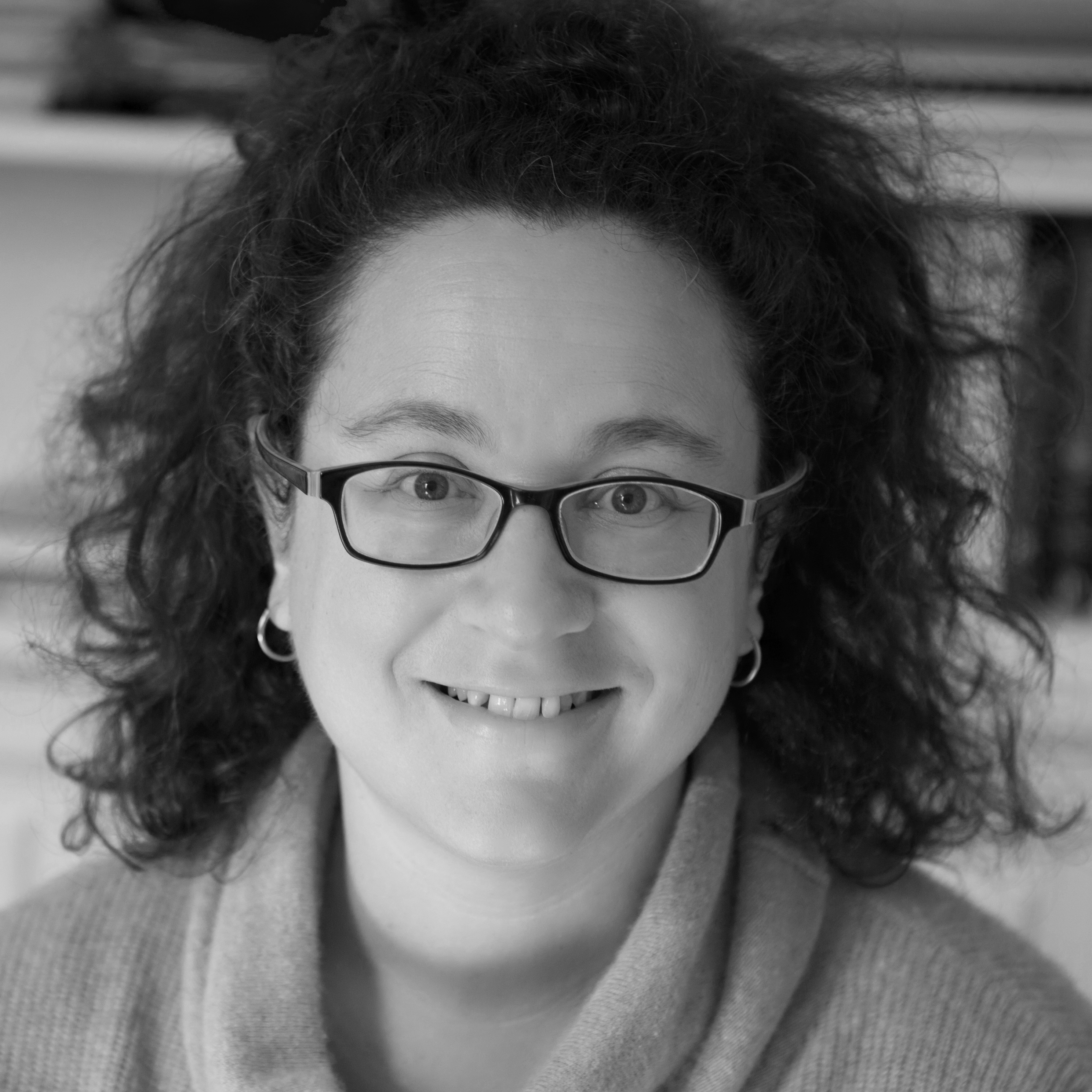 Black and white photographic portrait of Sophie Hannah who is smiling. She has dark wavy hair and glasses.
