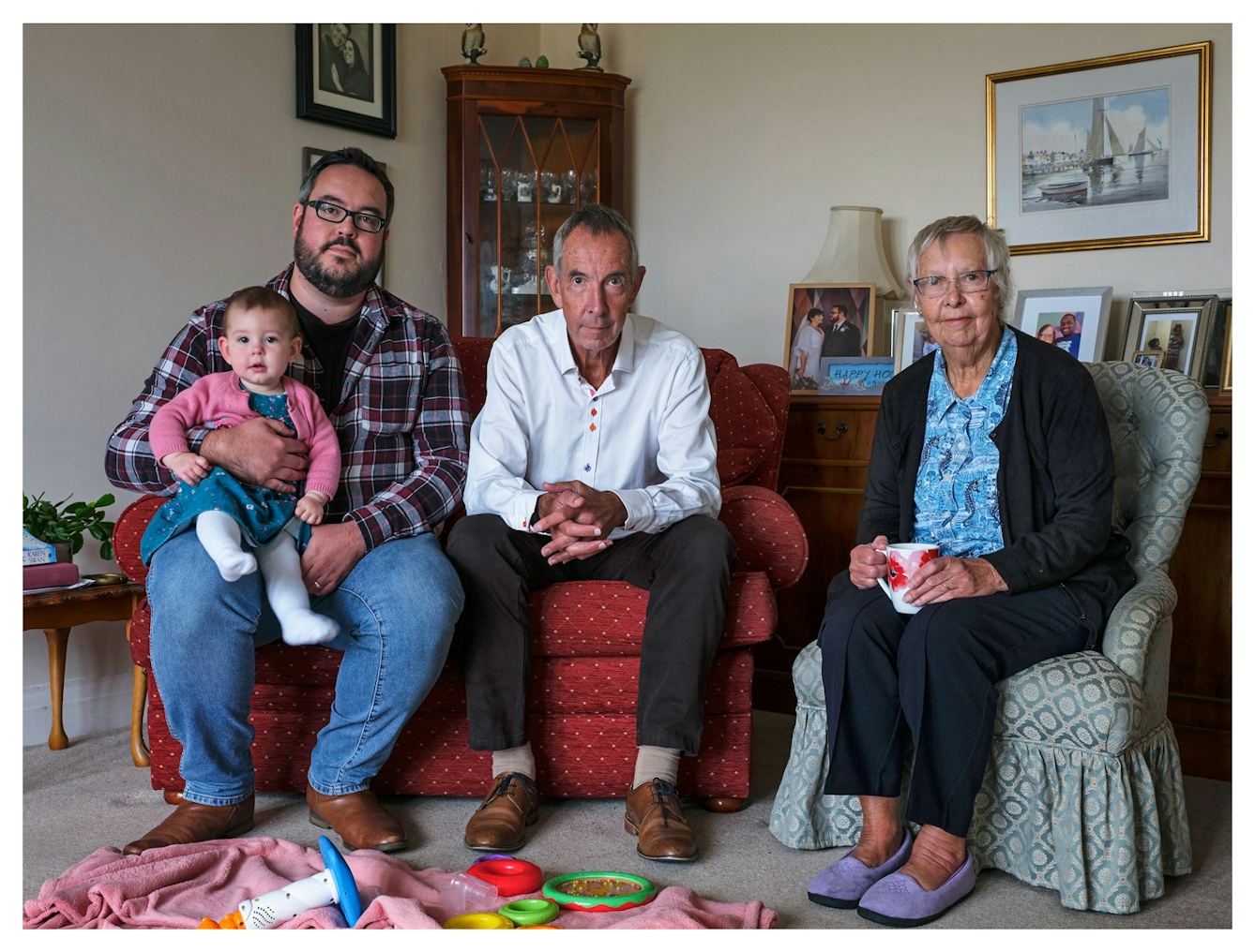 Family group photograph showing four generations of the same family. The great-grandmother sits on the right holding a hot drink in a mug, by her side sits the grandfather leaning forwards, fingers latticed. Next to him is the grandson who holds the great-granddaughter who is a toddler on his knee. They are all seated on a sofa and an armchair in a living room, surrounded by framed photographs, cupboards and a blanket on the floor covered in toddler toys. They all look straight into the camera.