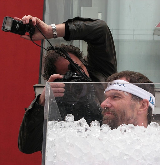 Photograph of Wim Hof submerged up to his head in a clear container of ice cubes. A photographer stood behind the container, holding a camera and light and taking a photo of Hof. 