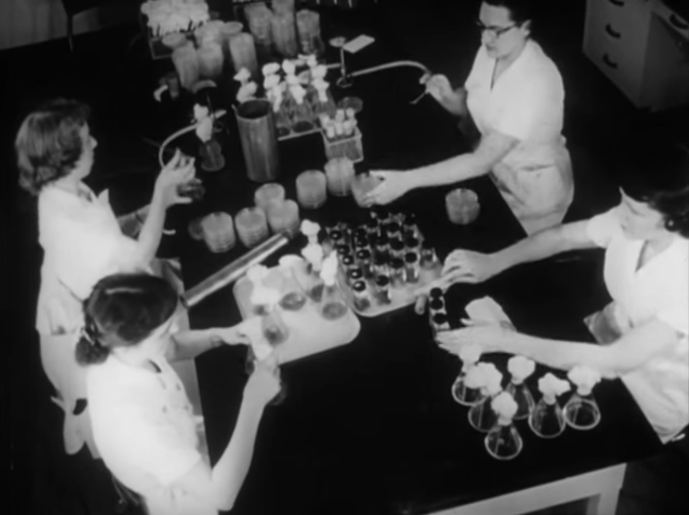 Still from back and white film featuring four women sitting around a lab table operating small cylindrical containers.