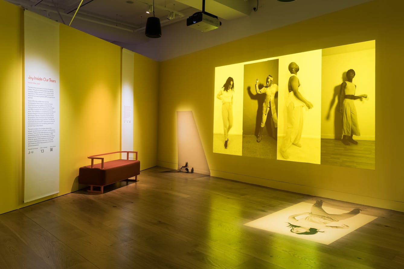 Photograph of an exhibition gallery space showing two walls meeting at right angles and a wooden floor. One the left hand wall is an information panel and bench seat. On the right hand wall is a projection of four large vertical photographs placed side to side, each monotone, but with a bright yellow tone overlaid. Each image shows an individual in a performative pose. The overall tones of the projection and the gallery walls are yellows.
