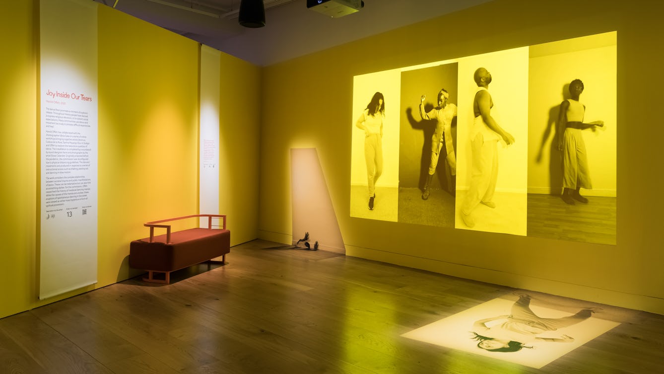 Photograph of an exhibition gallery space showing two walls meeting at right angles and a wooden floor. One the left hand wall is an information panel and bench seat. On the right hand wall is a projection of four large vertical photographs placed side to side, each monotone, but with a bright yellow tone overlaid. Each image shows an individual in a performative pose. The overall tones of the projection and the gallery walls are yellows.
