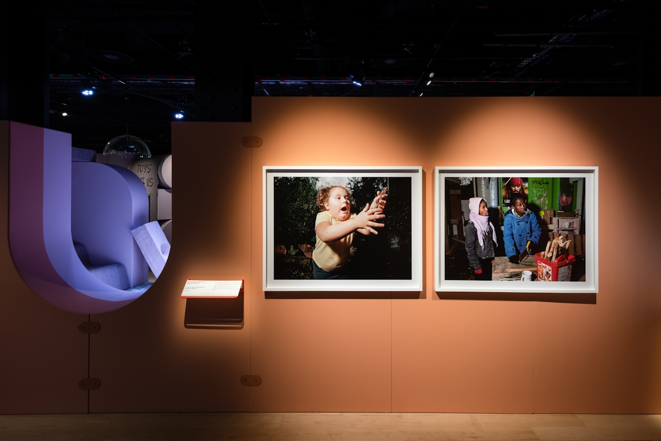 Photograph of an orange coloured exhibition wall showing 2 framed photographs. The photograph on the left shows a young girl reaching out to catch a frog. The photograph on the right shows two young people dressed in winter clothes sawing a section of wood.