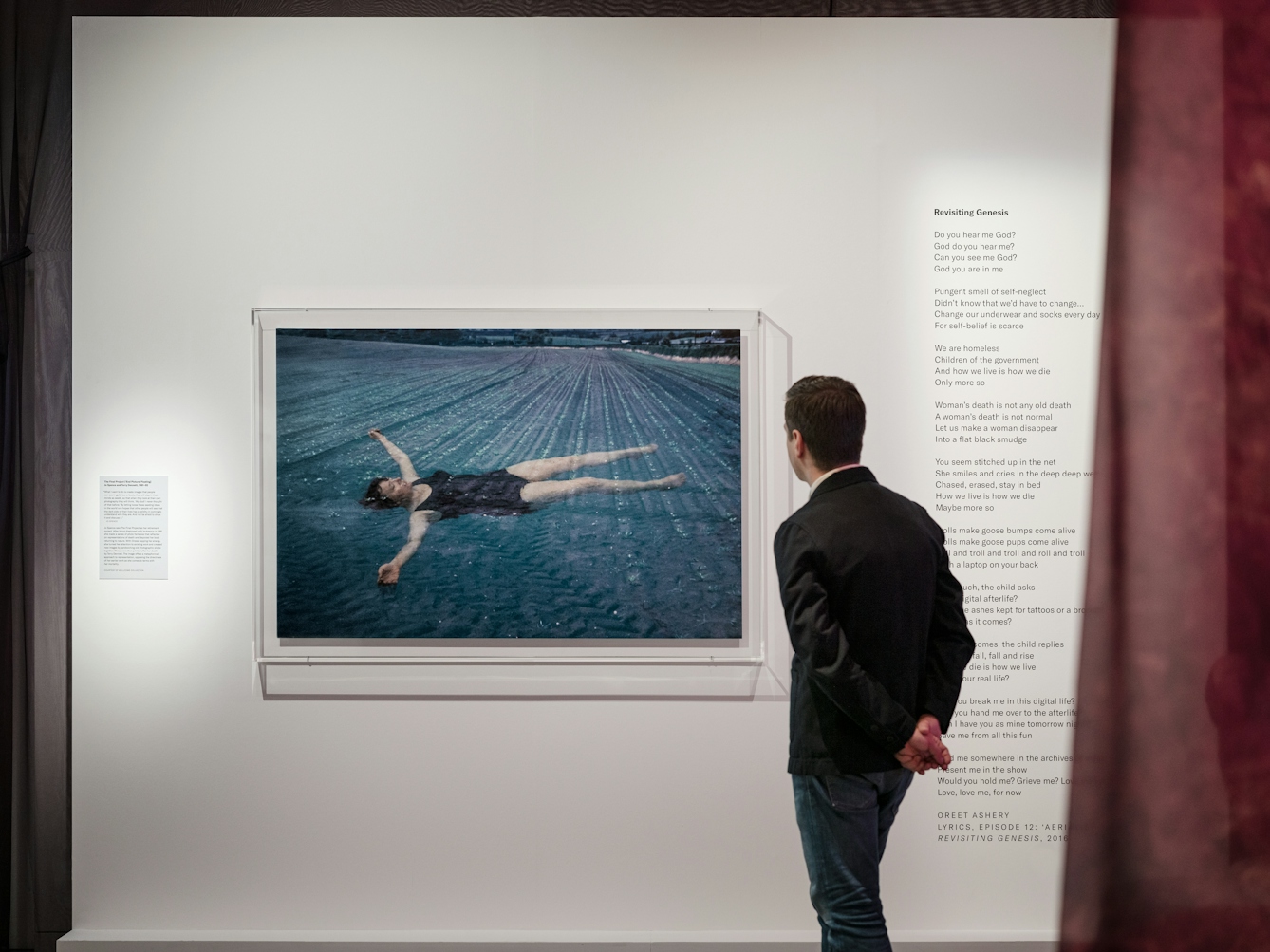 Photograph of a man in a dark jacket and jeans viewing a photograph of a woman floating belly-upwards in a blue pool.  To the right of the man there is a poem called Revisiting Genesis
