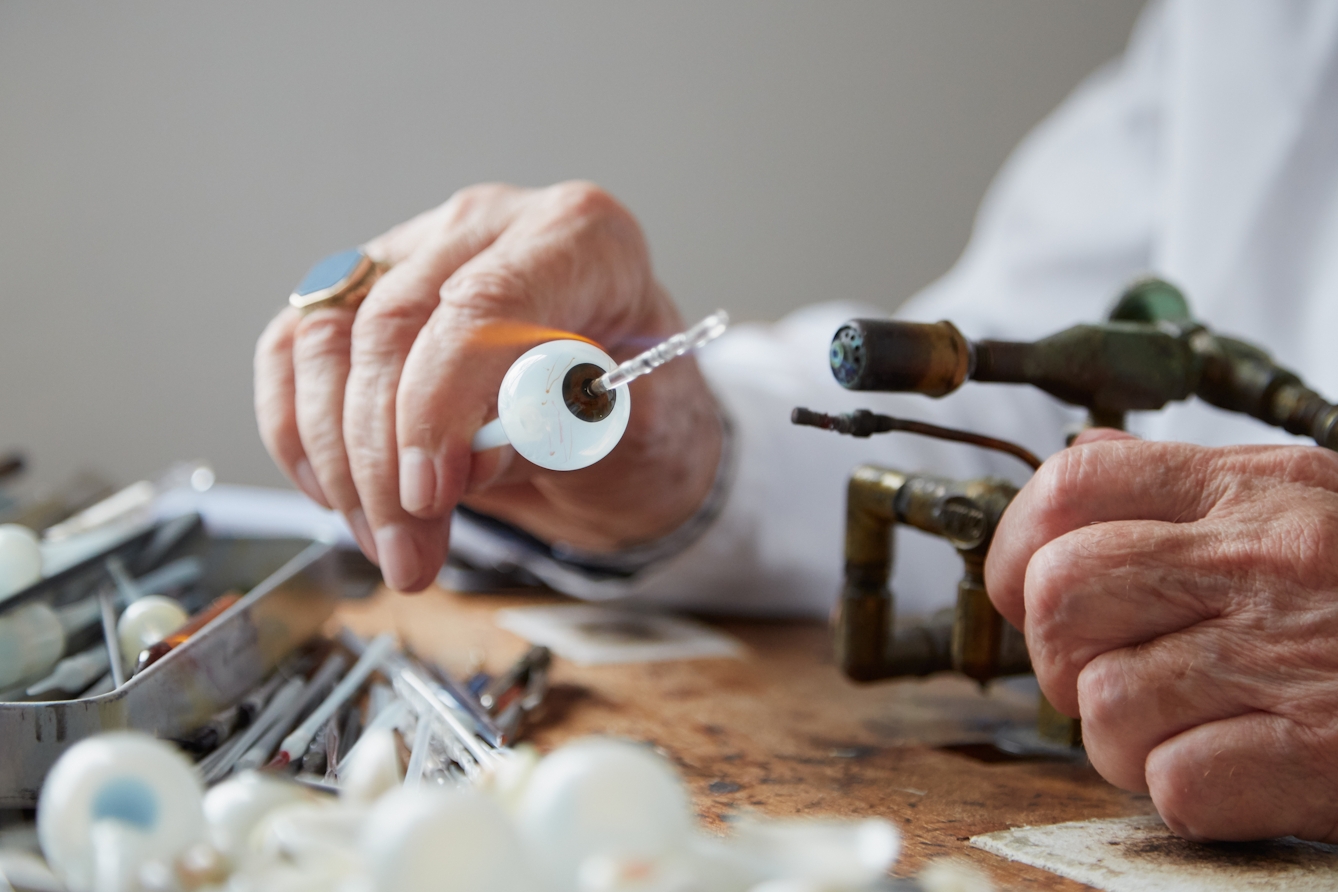 Photograph of the hands of a man in a white lab coat, holding a tube of white glass with a sphere resembling an eye ball at one end, to the flame of a gas torch. Next to him on the workbench are thin tools and more lengths of glass tubing.