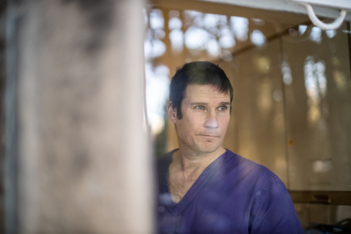 Photograph of a man from the chest up wearing blue medical scrubs. The photograph has been taken through a glass window and the man is gazing out of the window off to camera right, lips pursed.  Reflected in the window can be seen the out of focus blur of trees and sky. To the right is the also out of focus stone edge of the building.