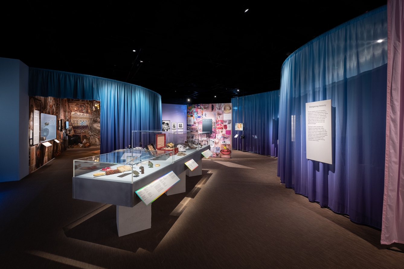 An exhibition space showing objects in glass-toped table displays, images and information boards on straight wall panels and suspended infront of curved floor to ceiling curtained walls.
