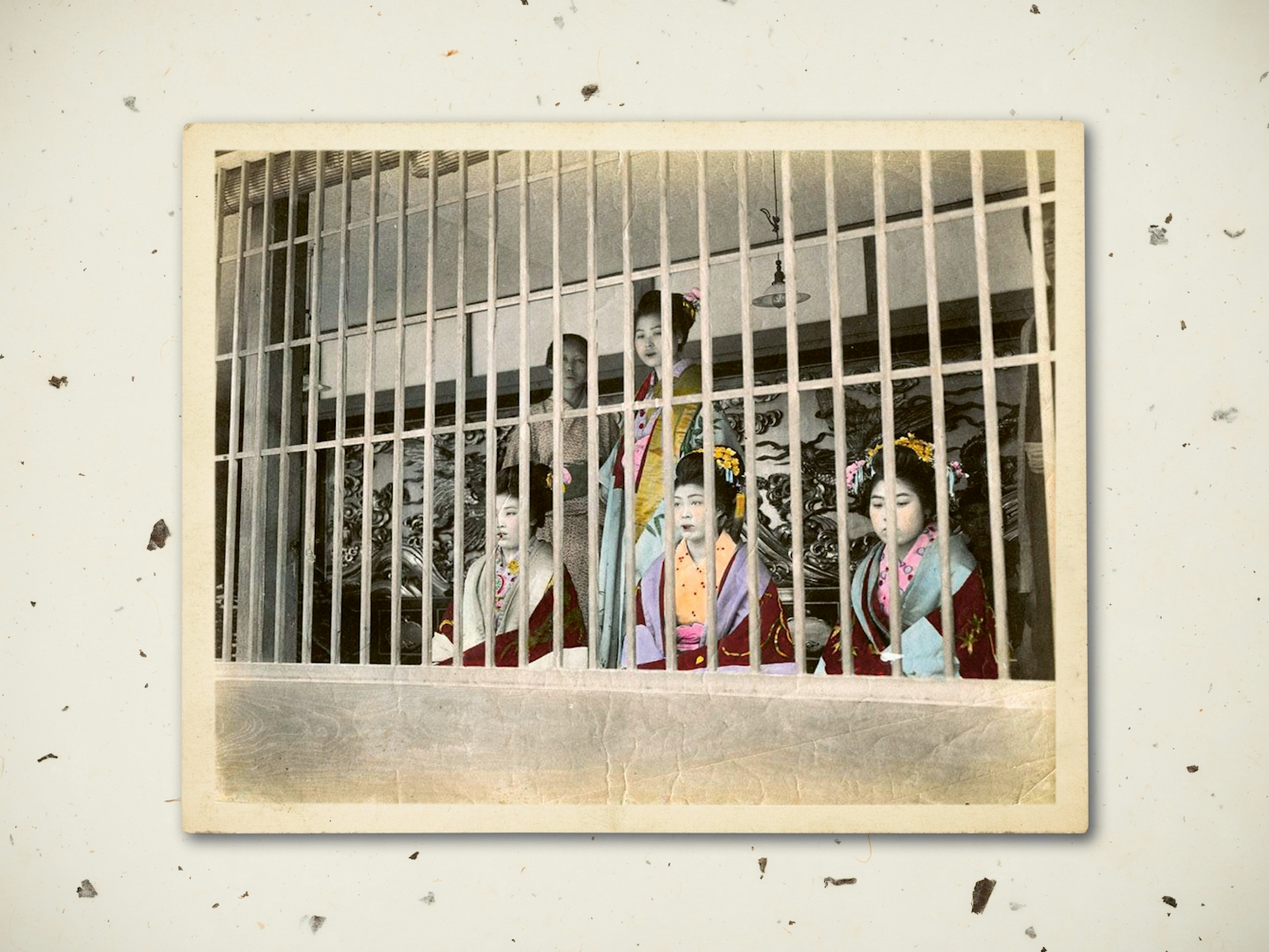 Digital composite image showing a textured rice paper background. Resting on top of the background is an early 20th century black and white photograph showing sex workers on display in the Yoshiwara district of Tokyo. They are pictured seated and standing behind a bared window. Their clothes have been hand tinted. Their expressions are blank and sombre. 