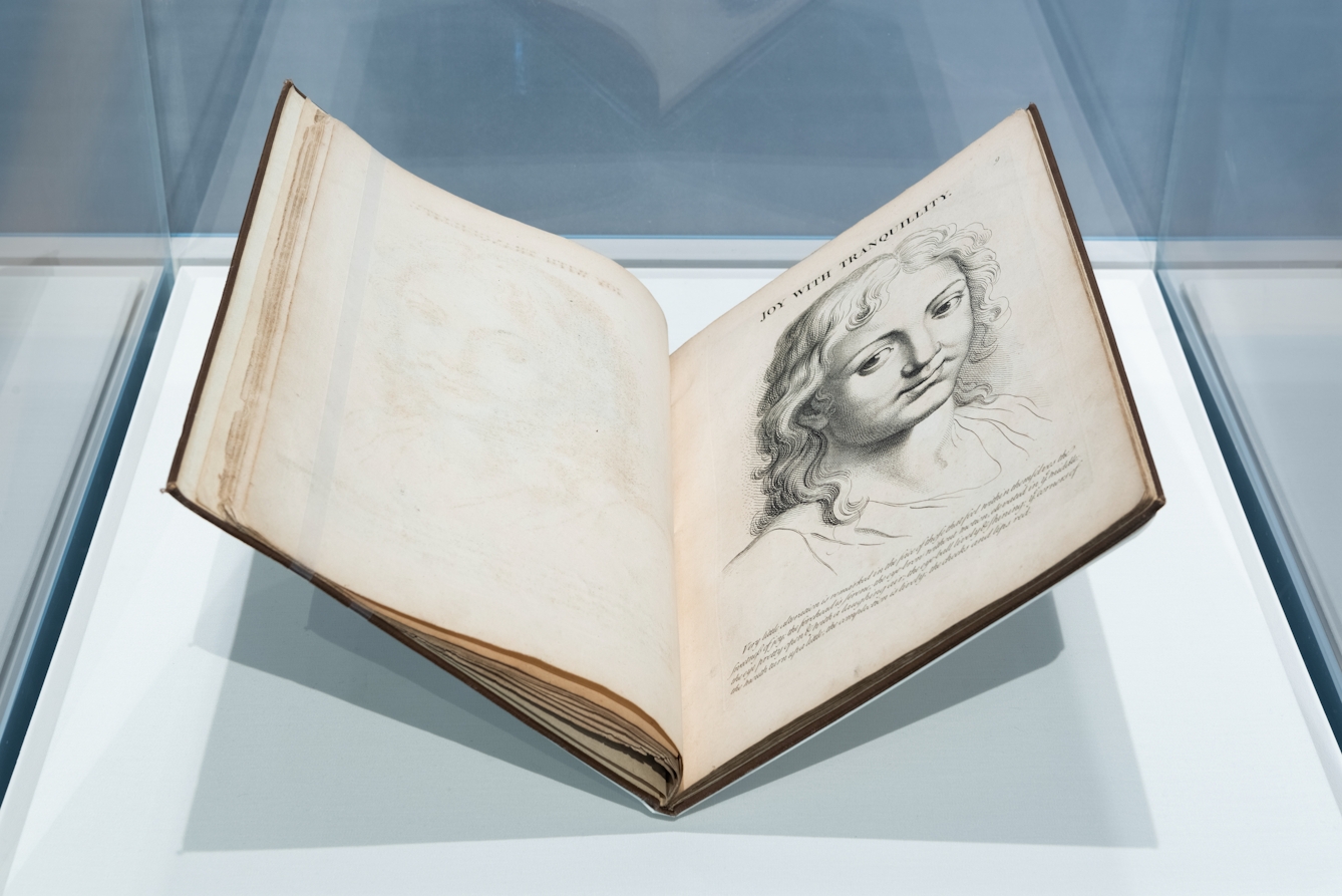 Photograph of an exhibition glass display case showing an open old, early printed book, held in a book cradle. The open double page spread shows a portrait engraving of a person's head with the title, 'Joy with Tranquillity' above it.