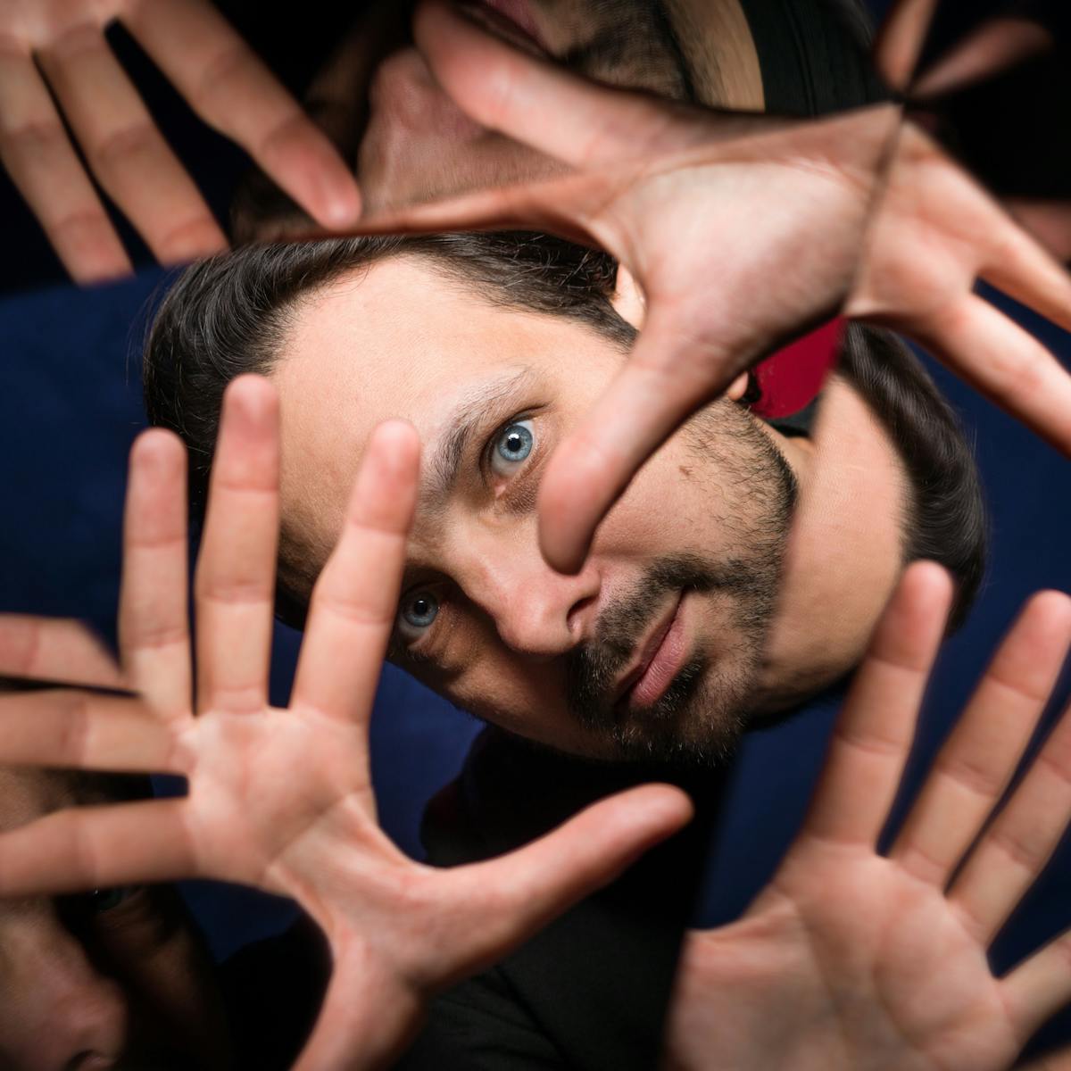 Photograph through a kaleidescope of the head of a man. His hands are held up in front of his face, with fingers outstretched, framing has eye. The background is made up of blue and red fabric.