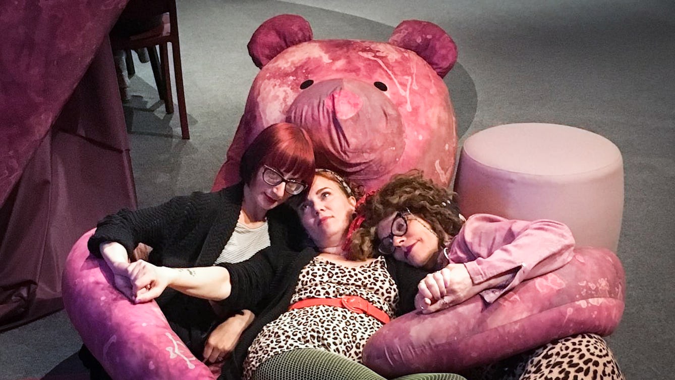 The artist Amanda Millis holding hands with two people on the floor lying down in the arms of a giant purple teddy bear in Wellcome Collection's Misbehaving Bodies Exhibition. 