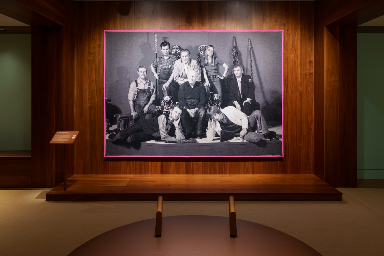 Photograph of a gallery exhibition space showing large tapestry hung on a wooden wall. The tapestry is made up of a black and white, archive photograph, from the late 20th century showing a group of white men and women gathered in a group within an exhibition exhibit. One of them is seated on an ornate throne with the others perched and lying around him, some holding museum artefacts in their hands. The tapestry is framed with a bright pink border. Surrounding the tapestry is a wider wooden structure containing seating and alcoves.
