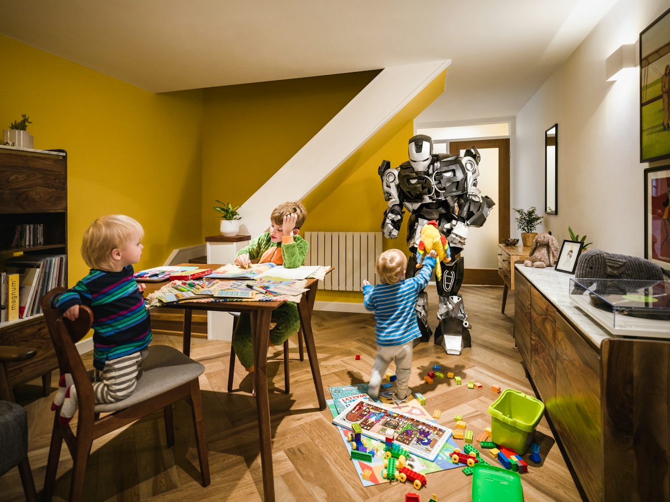 Photograph of a living room with two small toddlers and an older boy playing with puzzles, bricks and colouring books. In the background is a large robot bending down to receive a soft toy that now of the toddlers is handing it.