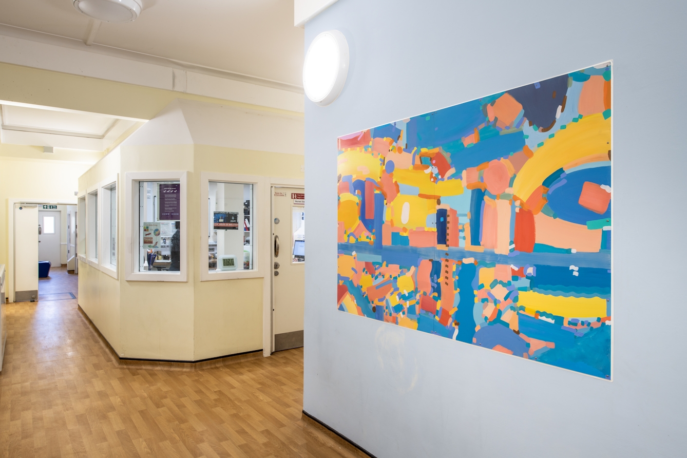 A photograph showing a corridor painted magnolia with wood laminate flooring. There is a high security internal office area surrounded by internal windows and a door. On the right-hand side of the image the wall is painted light blue and features a large rectangular canvas hung in landscape orientation. The canvas features a wide range of freely drawn irregular shapes in various tones of yellow, blue and orange. There are some straight, curved and even wavy lines. The abstract shapes resemble building, pathways, and rivers as though seen from a bird’s eye view. 