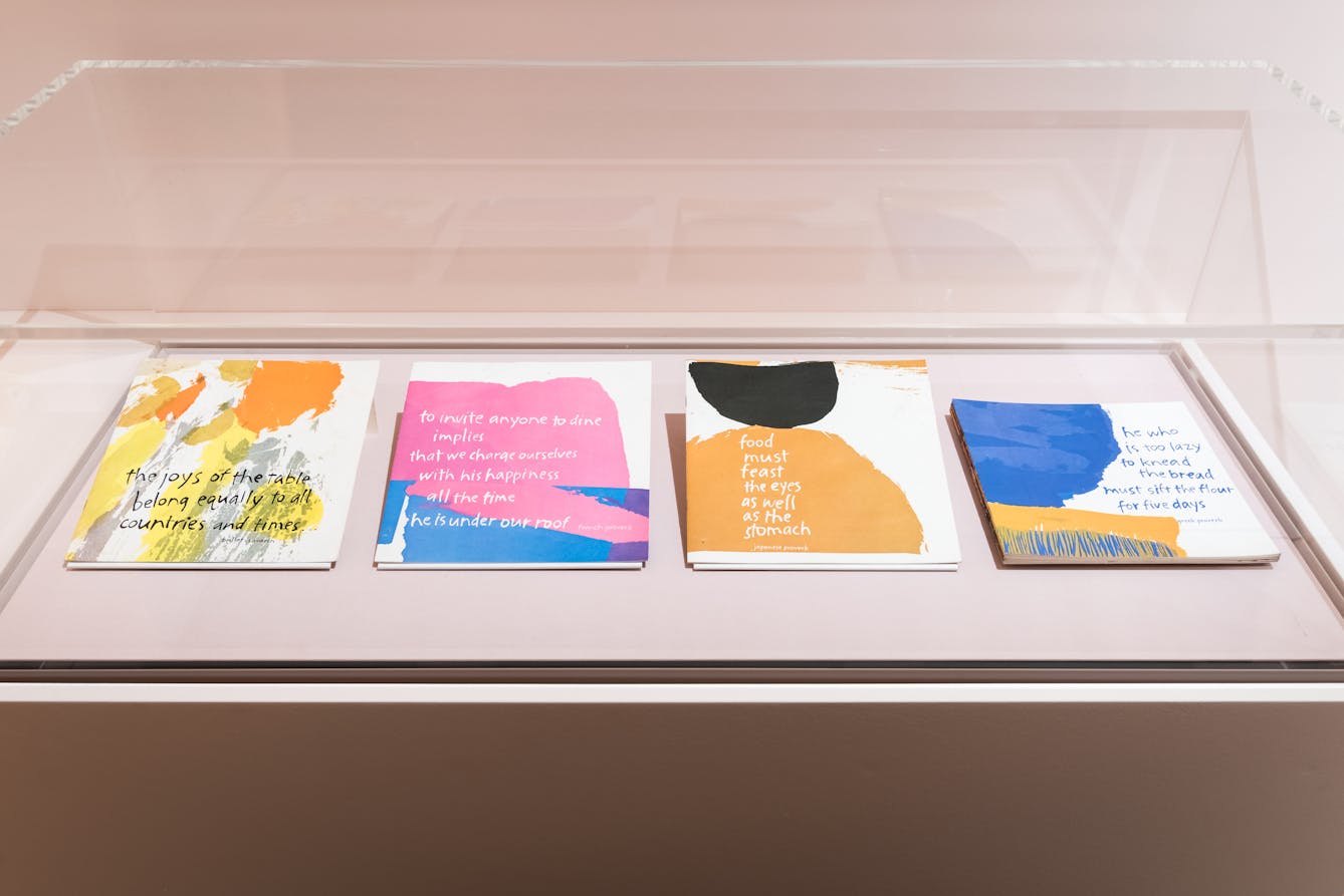 Photograph of a glass exhibition display case showing four colourful thin books laid out side by side, left to right. The covers contain text and abstract splodges of colourful shapes.