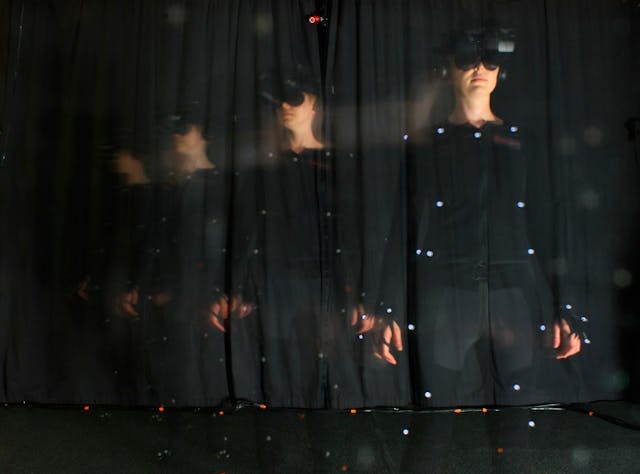 Colour photograph of a person wearing a headset over their eyes and black clothes against a black background. Their clothes have several dots over key places in order to track movements and match them in virtual reality.