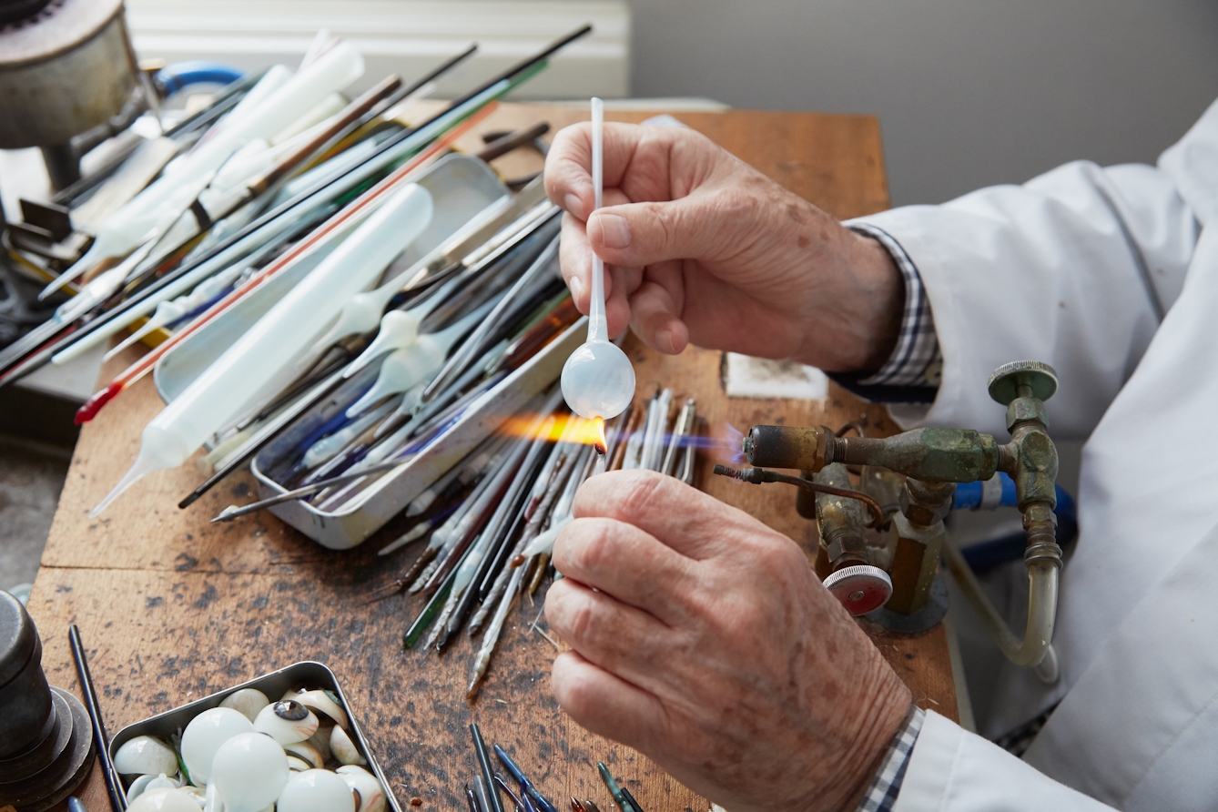 Photograph of the hands of a man in a white lab coat, holding a tube of white glass with a sphere at one end, to the flame of a gas torch. Next to him on the workbench are thin tools and more lengths of glass tubing.