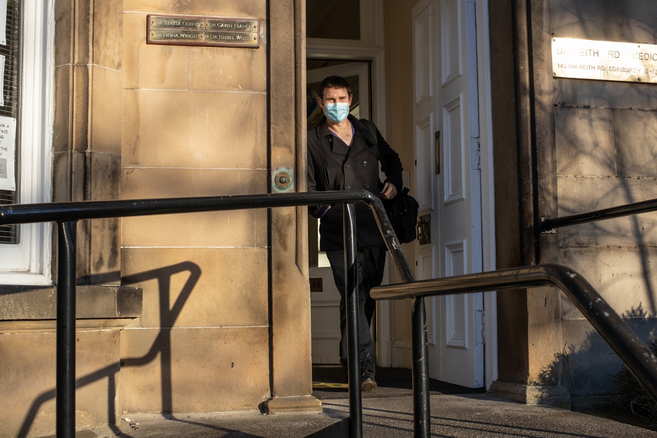 Photograph of a man wearing a black jacket and trousers and blue medical face mask walking out of the front door of a sandstone building. Over his left shoulder is a large black bag. In the foreground are the black handrails of the entrance steps. He is looking to camera as he walks. To the left and right of the doorway are brass plaques. The writing on the plaque to the right is obscured by reflected sunlight. The plaque on the left reads, 'Dr Teresa Quinn, Dr Gavin Francis, Dr Fiona Wright and Dr Ishbel White'.