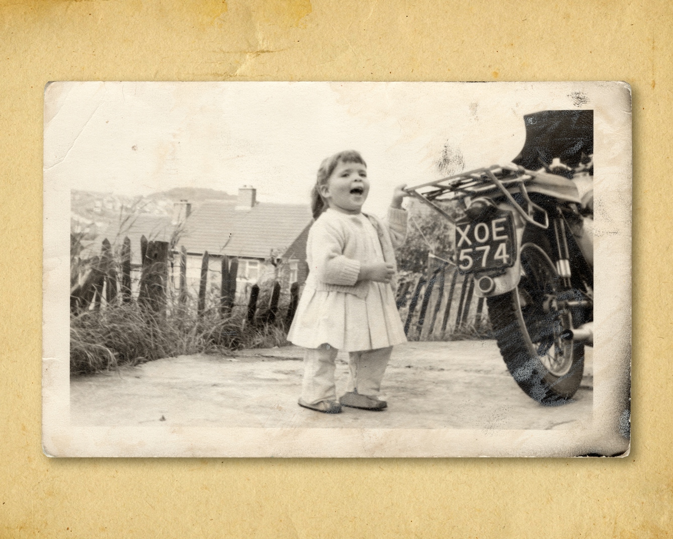 Photograph of a black and white photographic print, resting on a brown paper textured background. The print shows a small young girl in a dress, trousers and a cardigan standing outside. Her left hand is holding on to the pannier rack of a motorcycle which is just appearing into the frame. The motorbike has the registration plat 'XOE574'. The young girl has her mouth open as if mid shout. She has blocks of different sizes under her feet. Behind her in the background is a wooden fence, grasses and the roofs of houses in the distance.