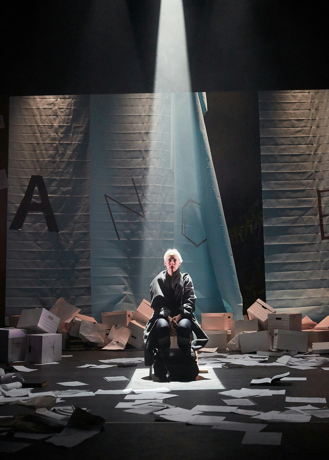 A woman sits ona  chair on s atge with spoltight beaming down on her from above. She’s surrounded by a mess of cardboard boxes and loose sheets of paper. 