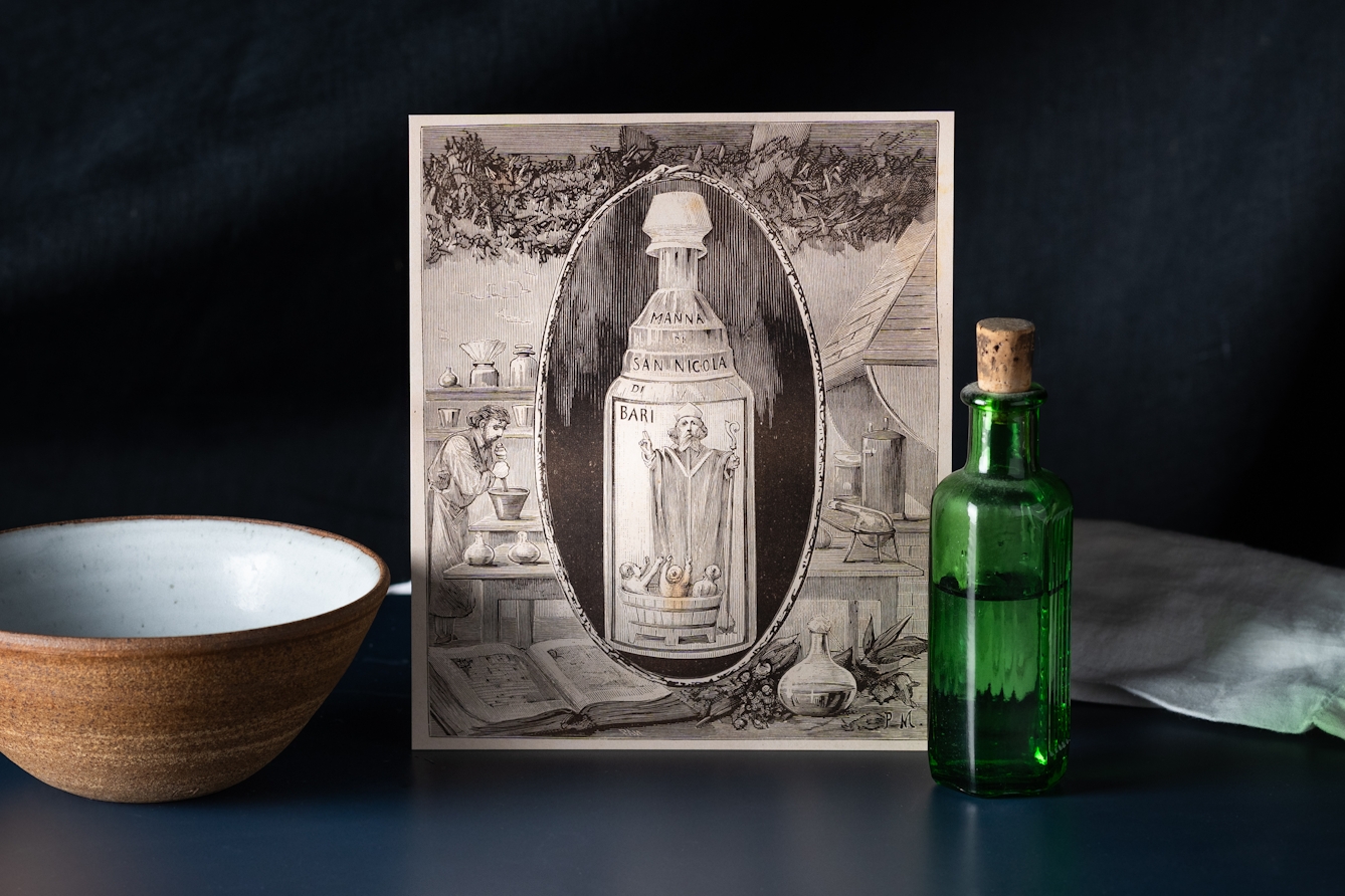 A still-life photograph showing a reproduction of an engraving depicting a bottle of the poison known as Aqua Tofana. The centre of the engraving shows a glass bottle with an illustrated label of a priest giving the sign of a blessing over three children in a wooden bath. Surrounding the bottle is a workshop scene showing a man surrounded by glass bottles and books who is mixing ingredients in a large bowl. Around the engraving is a stoneware bowl and a dark green apothecary bottle with a small cork stopper.