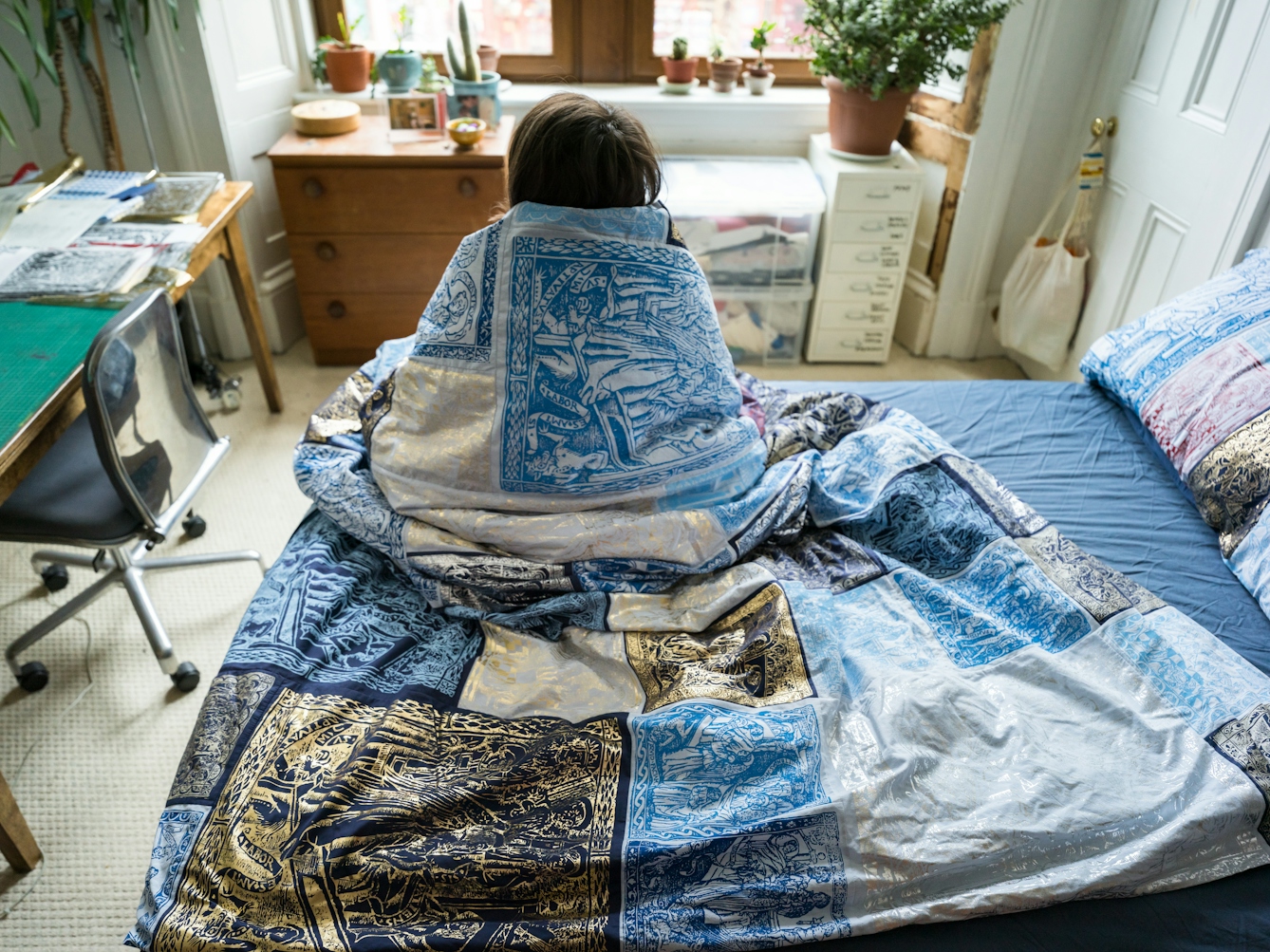 Photograph of a young woman sitting with her back to the camera on her bed, wrapped in a duvet cover made of a patchwork of blue, red, silver and gold screen-printed designs. She is looking towards large windows, a chest of drawers, a pot plant and the corner of a desk.
