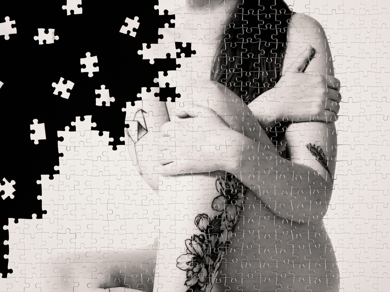 Photograph of a part made jigsaw puzzle containing hundreds of pieces. The jigsaw has been photographed against a black background, with many of the unused pieces scattered around in the black space. The image on the jigsaw is a warmed toned, monotone photograph of a nude woman against a plain light background. The woman is seated on the floor, pictured from the thigh to the neck. On her arm and leg are floral tattoos. The missing jigsaw pieces create a black void which encroaches from the left side of the image, fragmenting the image of the woman.