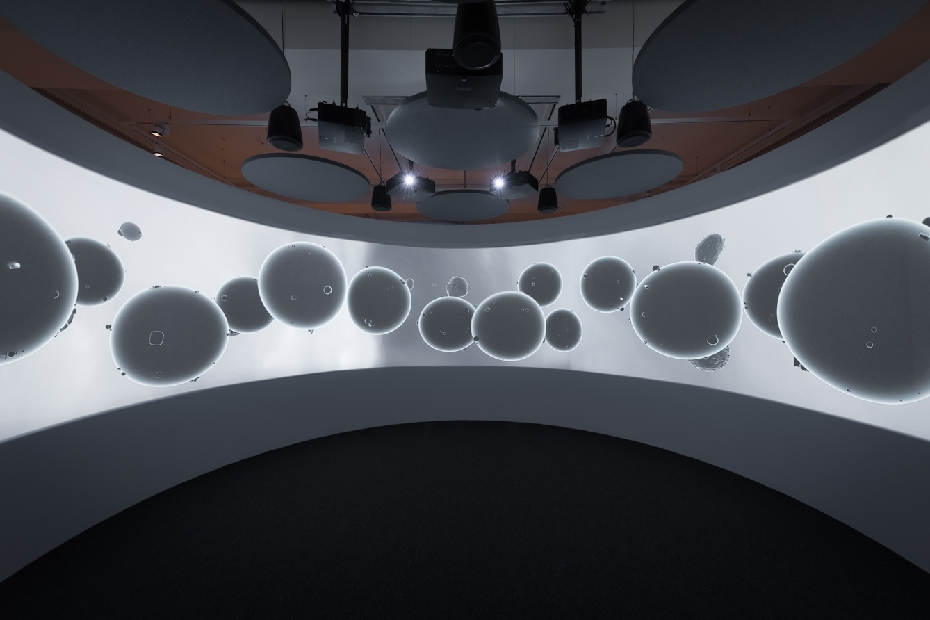 Colour photograph of a dark room with a very large, curved screen. Playing on the screen are about a dozen grey, particle-shaped round floating objects.  