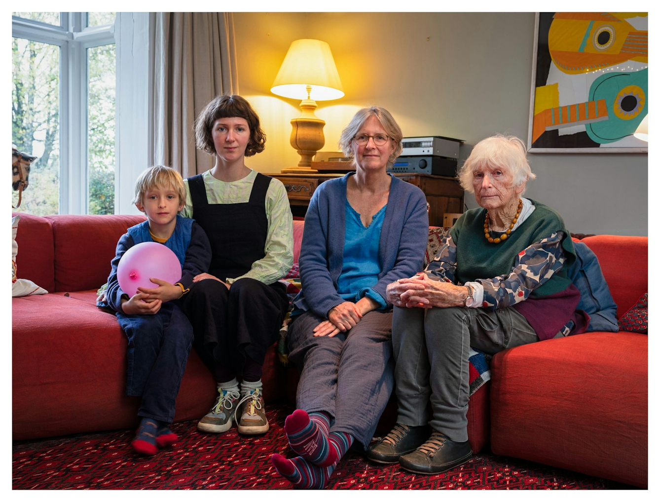 Family group photograph showing four generations of the same family. The great-grandmother sits on the right, by her side sits the grandmother. Next to her sits the granddaughter and next to her the great-grandson who is holding an inflated pink balloon. They are all seated on a long red sofa in a living room, surrounded by lamps, a sideboard and a framed picture. They all look straight into the camera.