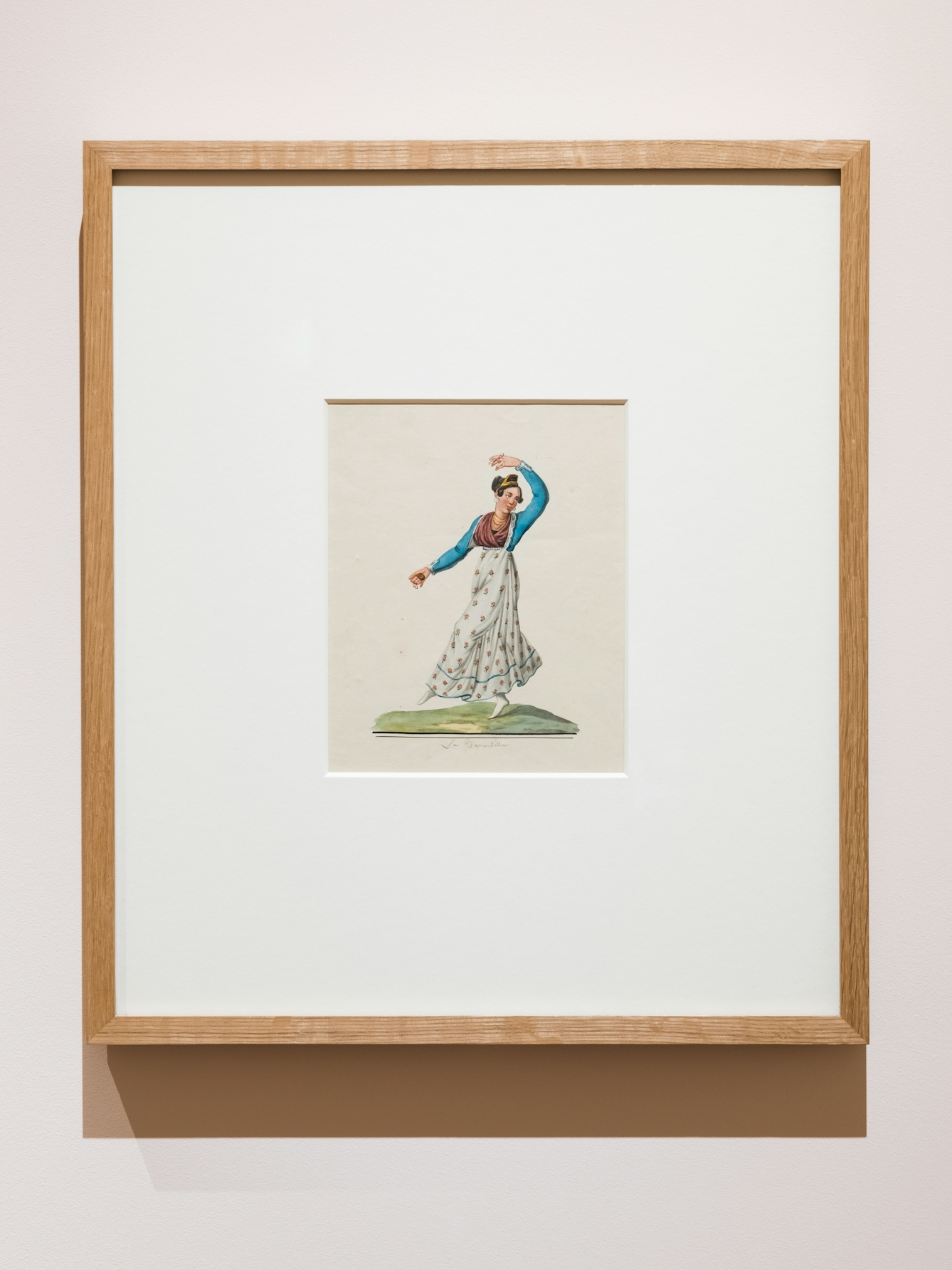 Photograph of an exhibition gallery showing a section of light pink coloured wall on which a framed print in a wooden frame has been hung. In a window mount within the frame is a colourful drawing of a woman dancing the tarantella, one arm raised over her head, one leg raised.