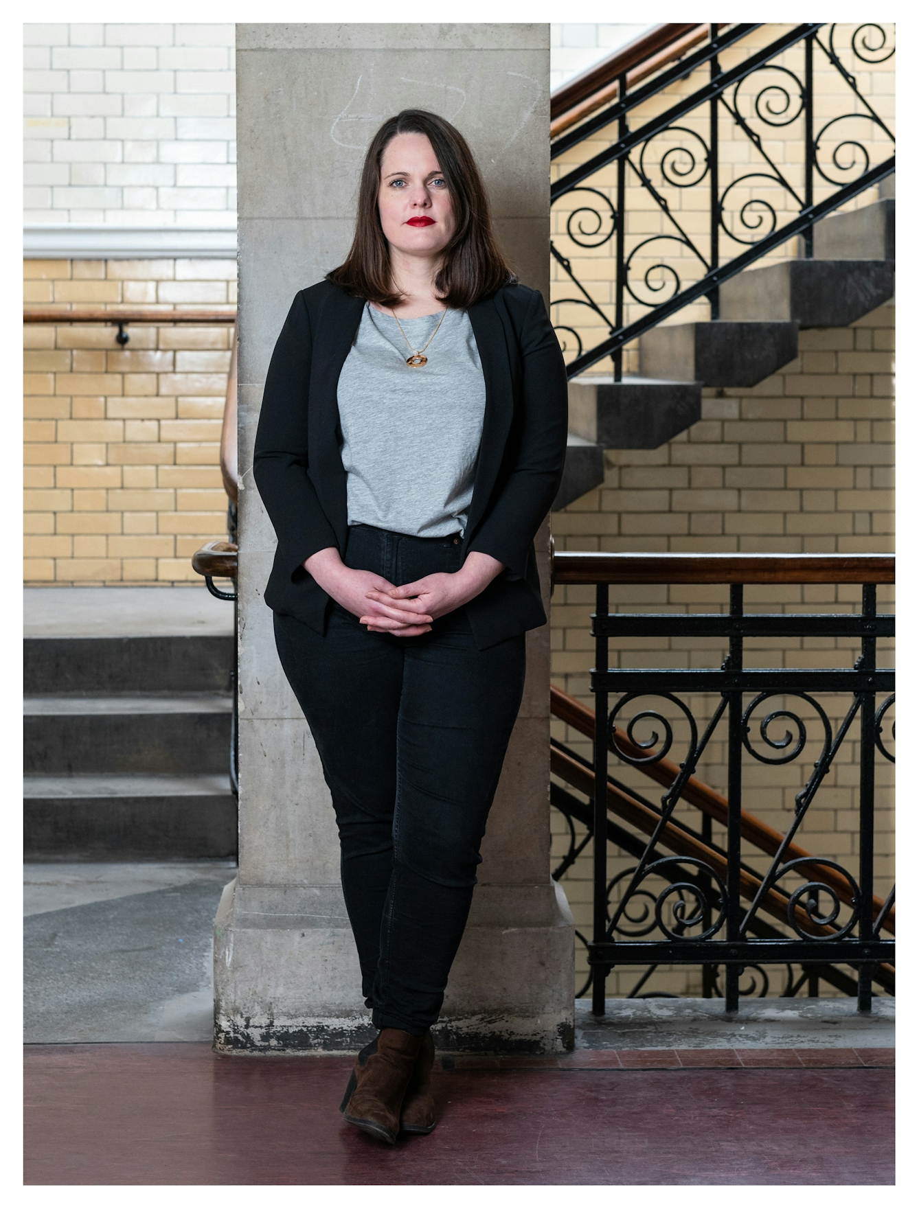 Photographic full length portrait of a woman standing looking to camera with her hands clasped in front of her black trousers. She is wearing a black long sleeved cardigan and with a grey top underneath. She is lit from the left by a soft light. Behind her are the glossy tiled walls of a Victoria period building and a large staircase with case iron balustrades made in intricate curls.