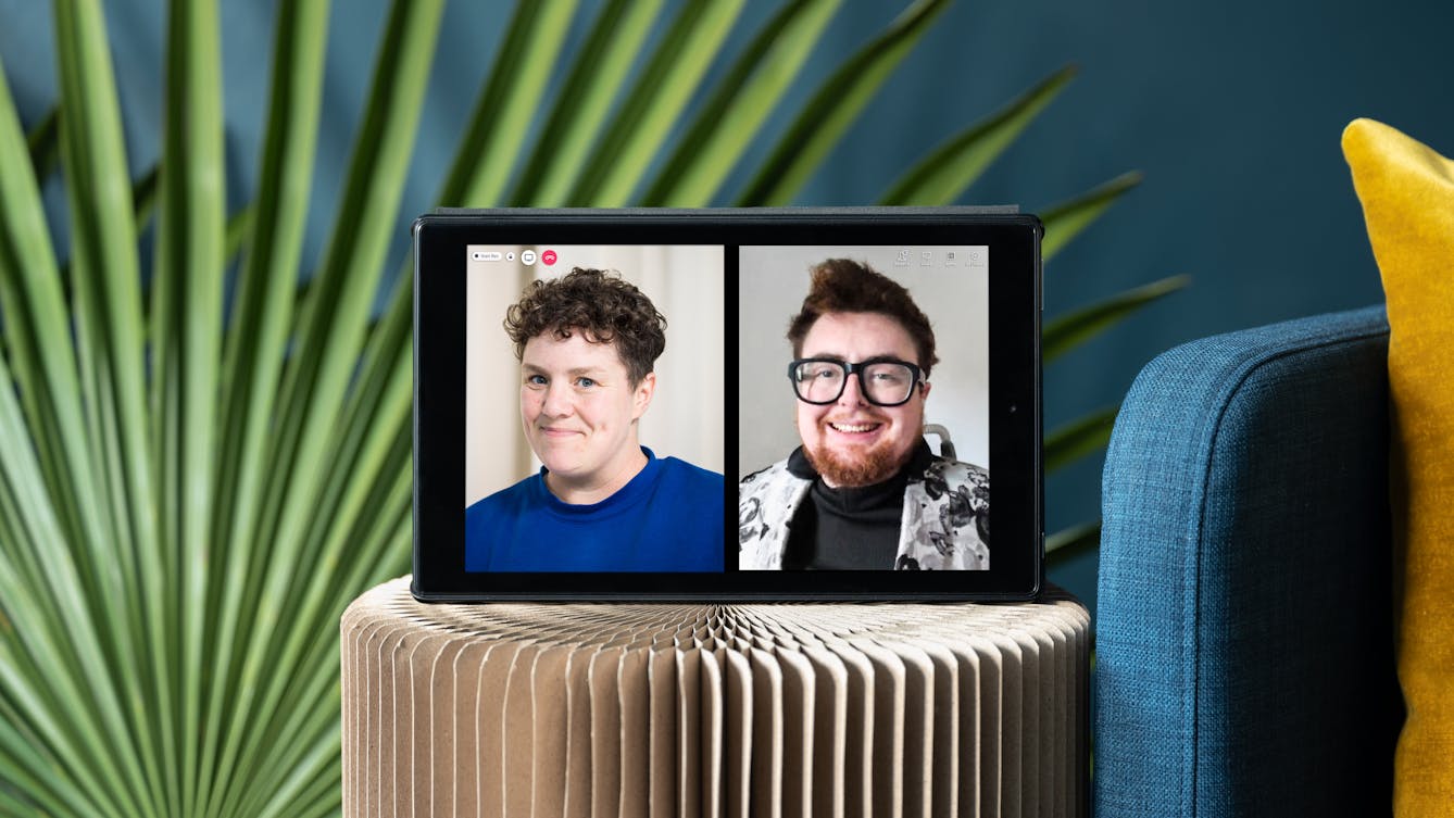 Photograph of a tablet standing on a stool. On the left half of the tablet screen is a photographic portrait of Jess Thom, wearing a blue sweater.  On the right half of the tablet screen is a photographic portrait of Jamie Hale, wearing glasses and a black sweater with a patterned jacket over it. Also, on the screen are video call icons "People", "Chat" and a red telephone icon. There are green decorative palm leaves displayed in the background behind the tablet which is next to the edge of a blue sofa with a yellow cushion.
