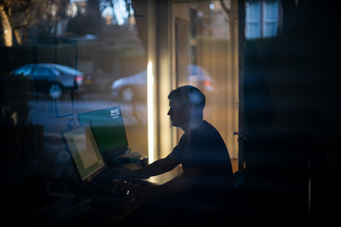 Photograph of a man through a glass window, sat at a desk in front of two computer screens. On the screen to his right are the words 'NHS Lothian'. The overall tone of the image is dark, with the man silhouetted against bright sunlight against the room behind him. Reflected in the window, the out of focus street scene can be seen; parked cars, house and trees.