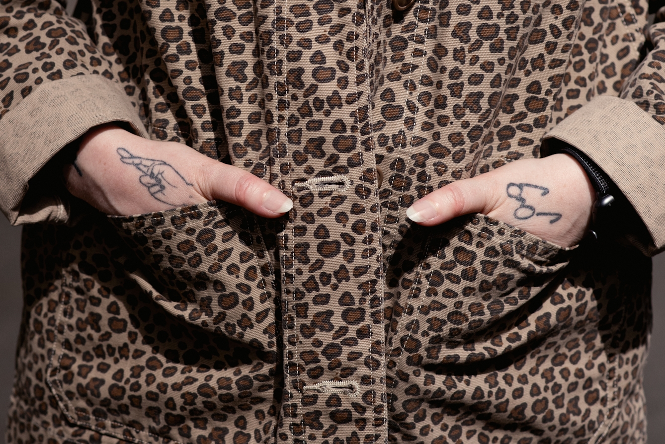 Close-up photograph of a woman's hands resting in the pockets of her jacket. Her thumbs remain outside the pockets. One her left hand is a small tattoo of a pair of spectacles. On her tight hand is a small tattoo of a hand with its fingers crossed. The jacket has a leopard skin pattern which fills the whole frame.