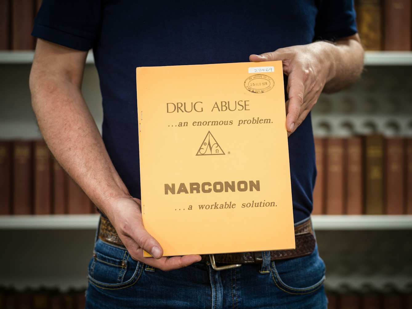 Photograph of the midriff of a man who is holding a pamphlet titled 'Drug abuse ...an enormous problem - Narconon ...a workable solution'. In the background are a set of out of focus shelves containing books.