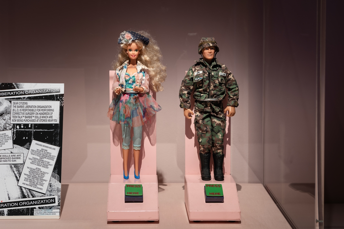 Photograph of an exhibition display case containing a male and a female doll. The female doll is dressed in floral outfit, the male doll is dressed in army fatigues. In front of each doll is a green button with the words, 'PRESS HERE' written on them in red.