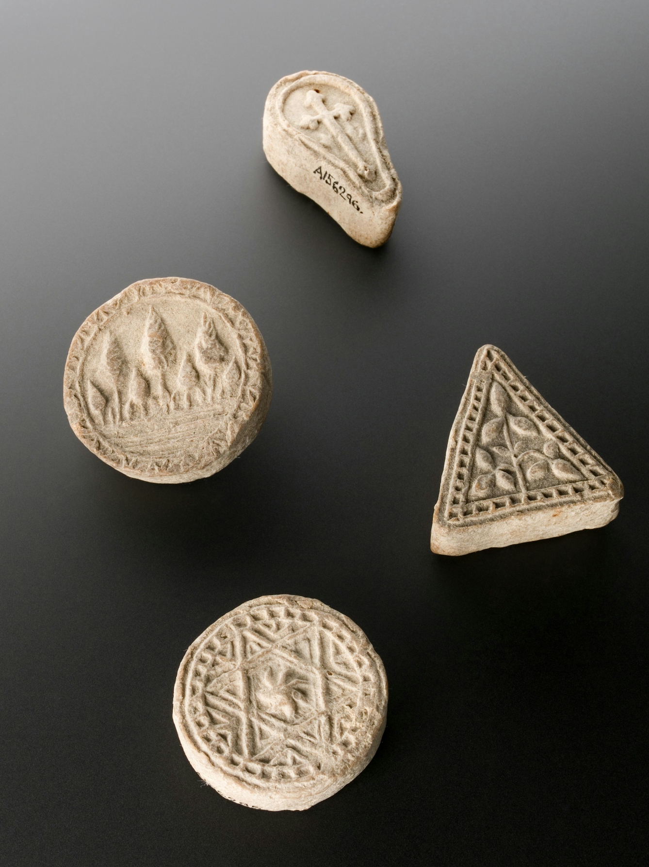 Photograph showing four shaped and decorated biscuits lying on top of a grey background. The top biscuit is an oval shape and has a Latin cross decoration. The second biscuit is circular and has a decoration of leaves on it. The third biscuit is triangular and has a similar leaf decoration. The fourth biscuit is circular and patterned with a star. 