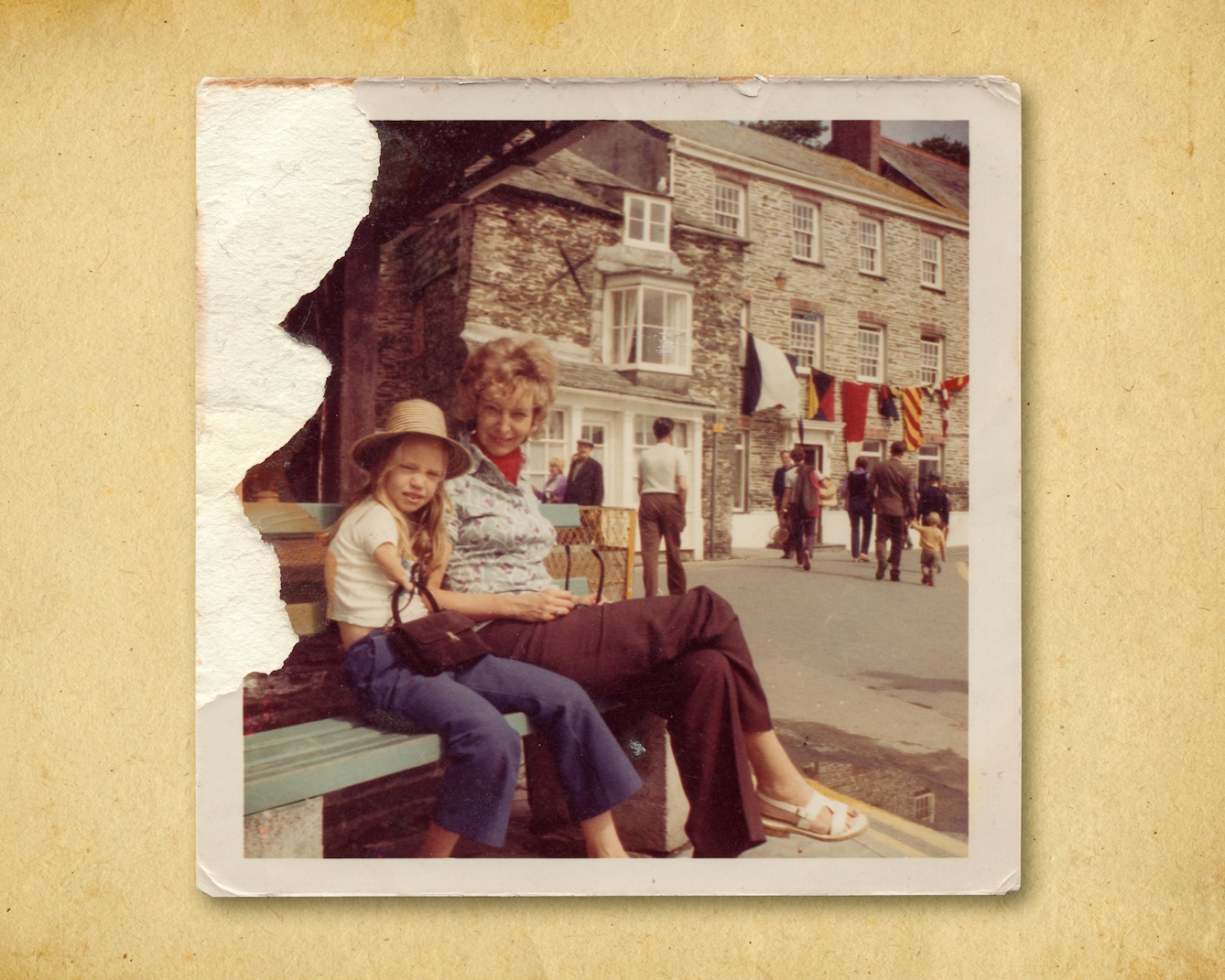 Photograph of a colour photographic print, resting on a brown paper textured background. The print shows a woman sitting on a public bench next to her young daughter. Behind them is large stone built building with a string of large naval flags hung across its facade. There are people in the distance walking toward the building. The mother and daughter on the bench are looking to camera. The daughter is wearing a summer brimmed hat and holding a hand bag in her right hand. The top left corner of the print has been torn such that the surface of the print and therefore the image have been lost.