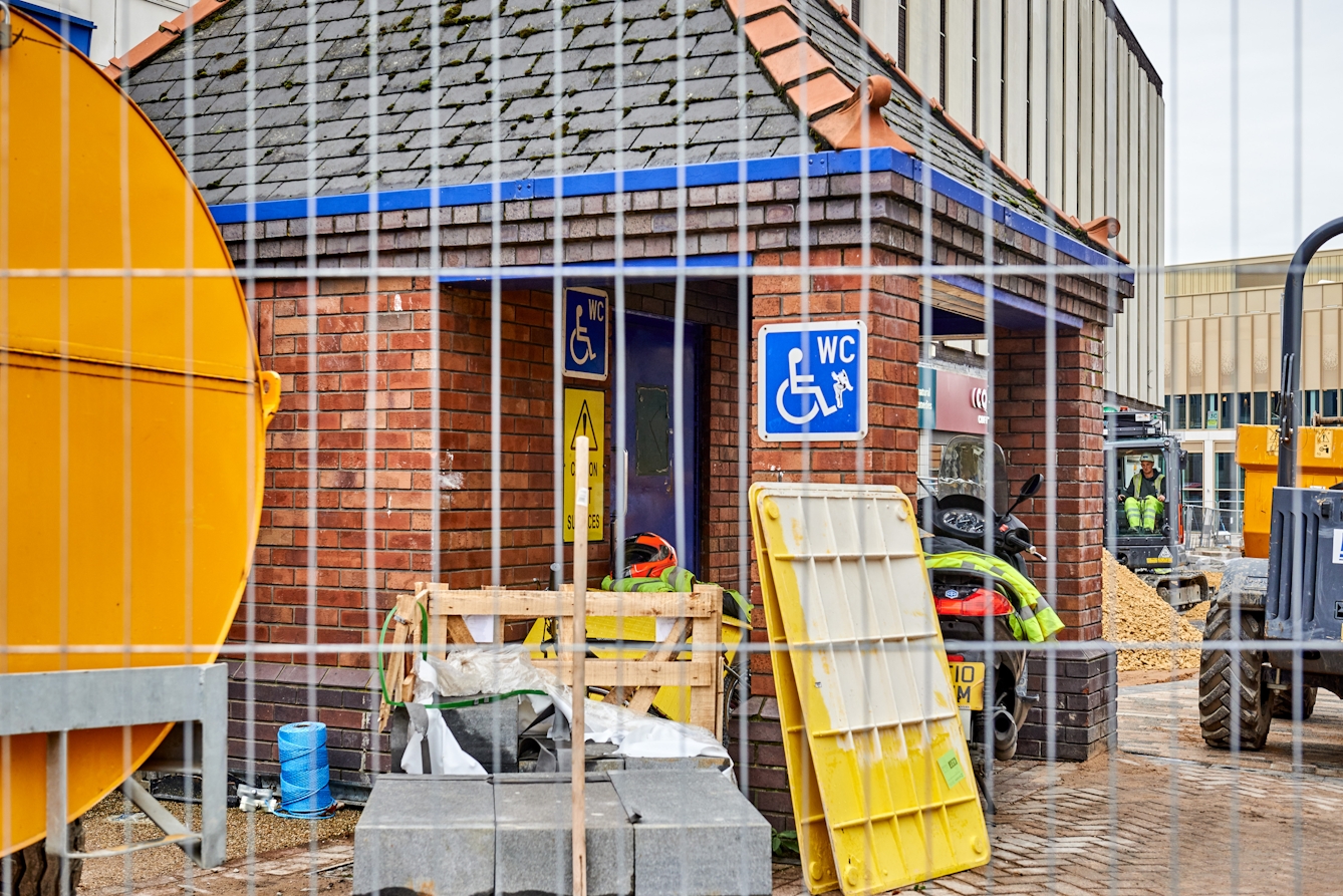Photograph of the exterior of an accessible toilet building, through a metal construction fence. The toilet is closed and the building is surrounded by construction materials and equipment.