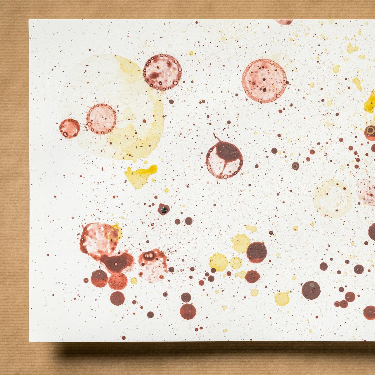 Photograph of an original artwork on watercolour paper. The artwork is resting on a brown parcel paper background. The artwork shows many varying in size droplet spatters and rings, made with coloured ink. The ink colours are dark red, yellow  and light red. The spatters seem random in distribution.