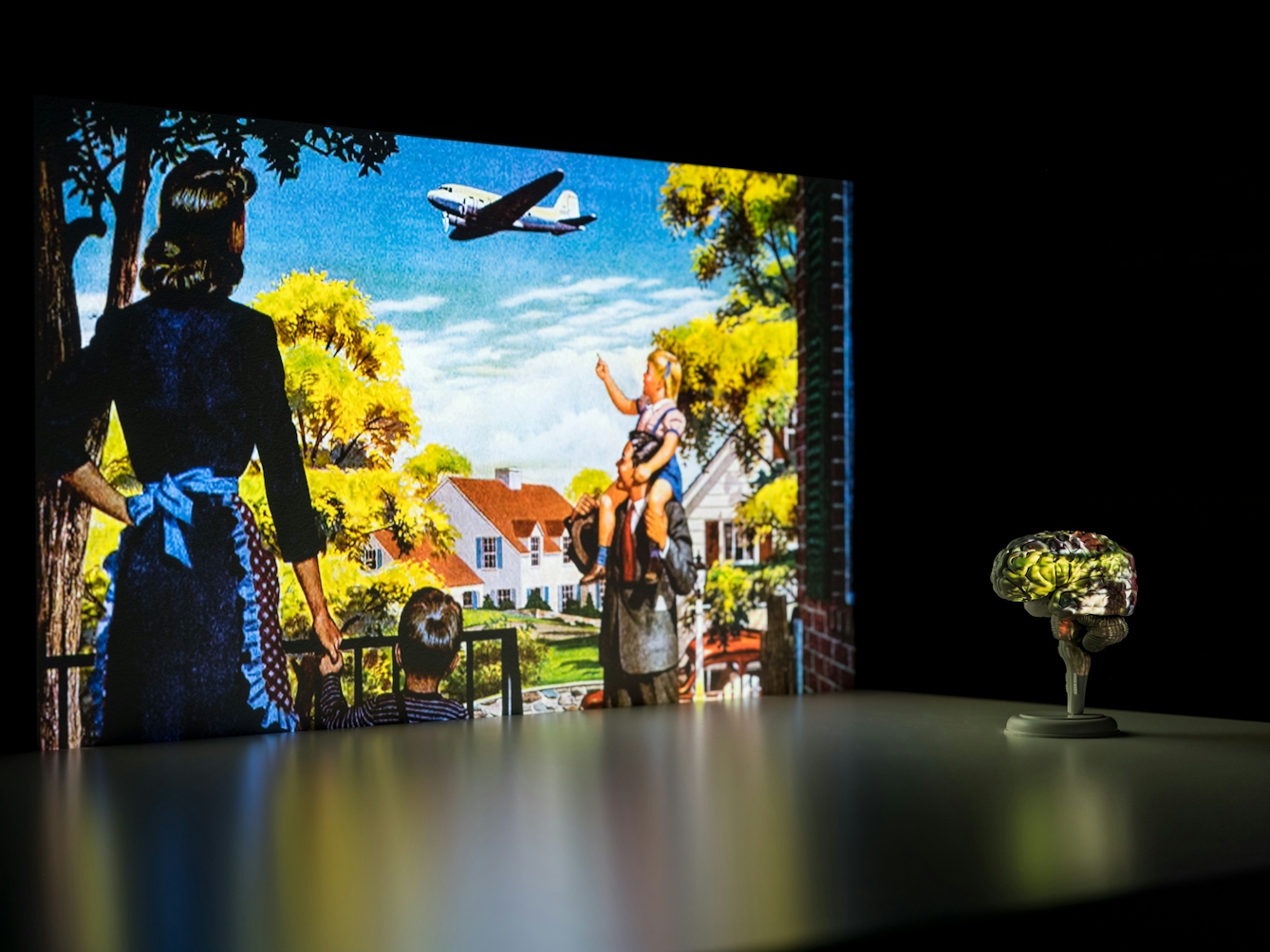 Photograph of a dark corner of a room. On the wall is a projected illustration from an American advertising poster. The scene shows a family group standing outside their suburban home looking up at an aeroplane flying past. In front of the projected image is a flat tabletop which shows a slight reflection of the illustration. Standing on the table to the right is a small model of a brain, resting on a stand. Part of the projected image washes across the surface of the brain, covering it in the image and its colours.