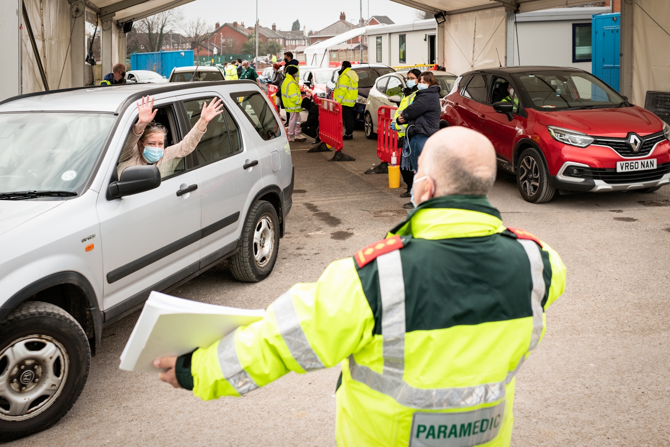 Photograph of a drive-through vaccination centre taking place in a car park. There are queuing cars waiting for the occupants' turn to received the vaccine. In the distance people are stood in hi-viz jackets. In the mid-ground to the left a car is driving away. The woman in the passenger seat has both arms stretched out of the window, arms held high in celebration. She is wearing a blue medical face covering. In the foreground, slightly out of focus, is the back of a man holding out a direction sign. He is wearing a hi-viz jacket with the word 'Paramedic' written on the back.