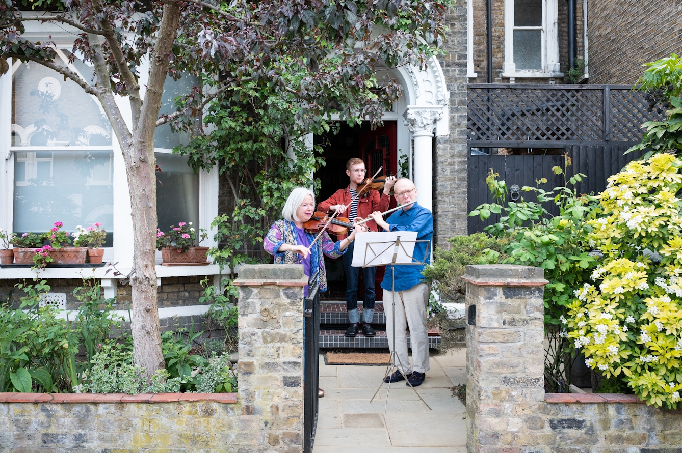 Photograph of the doorstep and front garden of a Victorian house. In front of the step and mother, father and son stand in front of a music stand playing the violin and flute.