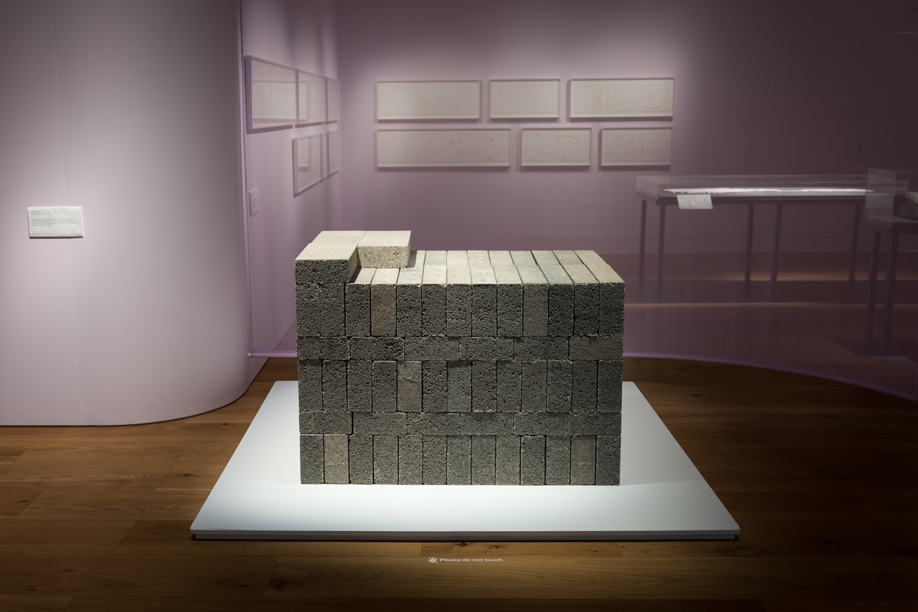 Colour photograph showing an exhibit in a museum. The exhibit is a large cuboid structure made of stacked rectangular grey and beige concrete blocks. The exhibit is on a white stand. There is a purple wall behind the exhibit with a number of framed images on it. To the left of the images there is a display table with a glass covering. 