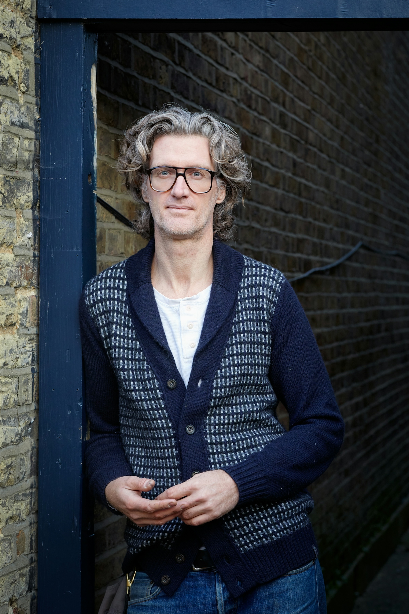 Photograph of a man wearing a patterned cardigan leaning against a blue doorframe attached to a brick wall. He is looking to camera and is wearing a large pair of thin rimmed spectacles. He has flowing wavy hair.