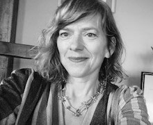 A black and white selfie photograph of a woman with shoulder-length fair hair. She is smiling and looking at the camera.